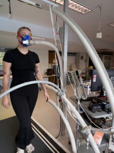 Picture of a female appearing person walking on a treadmill with a mask on their face.