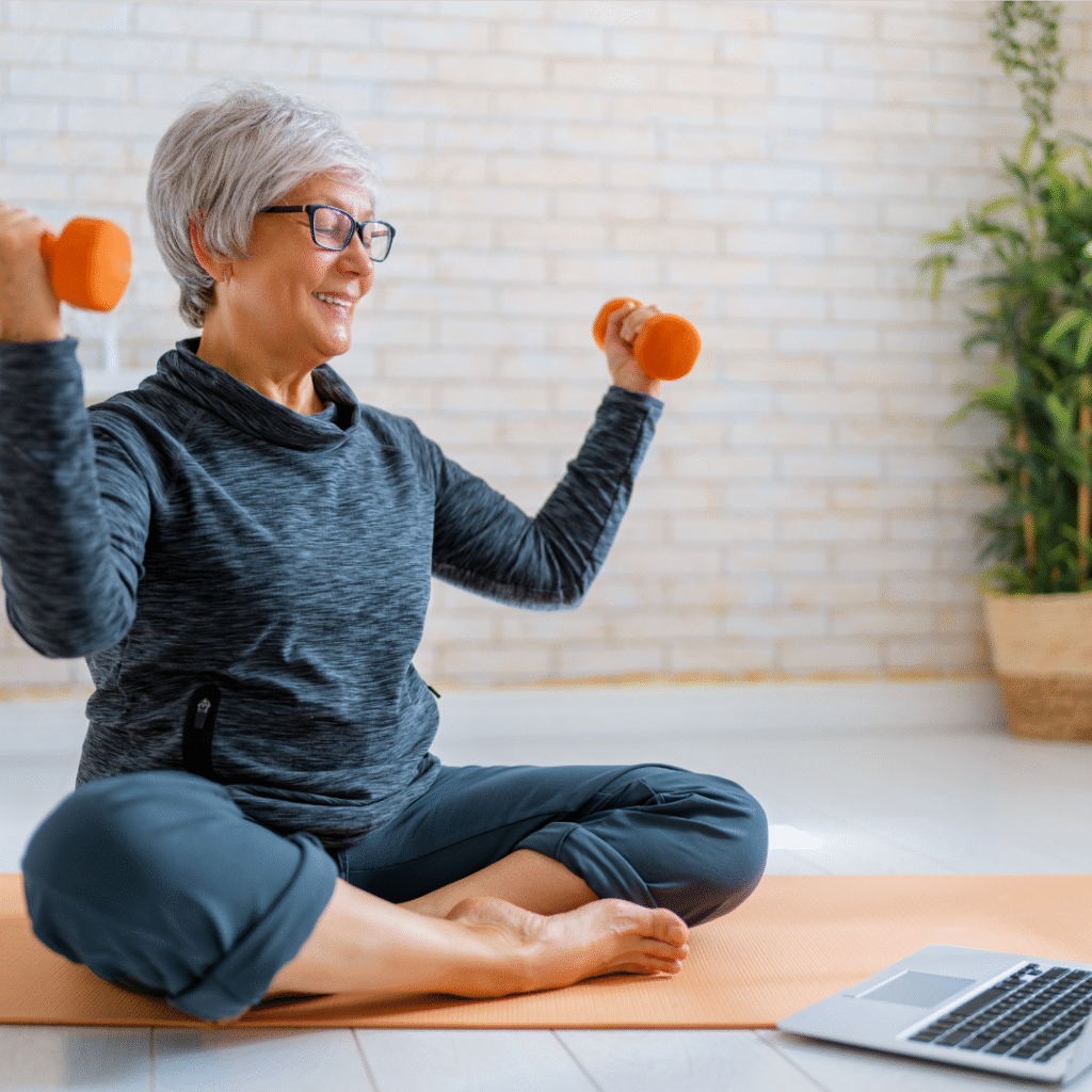 Female presenting adult holding weights in hand and sitting on gym mat looking at a computer