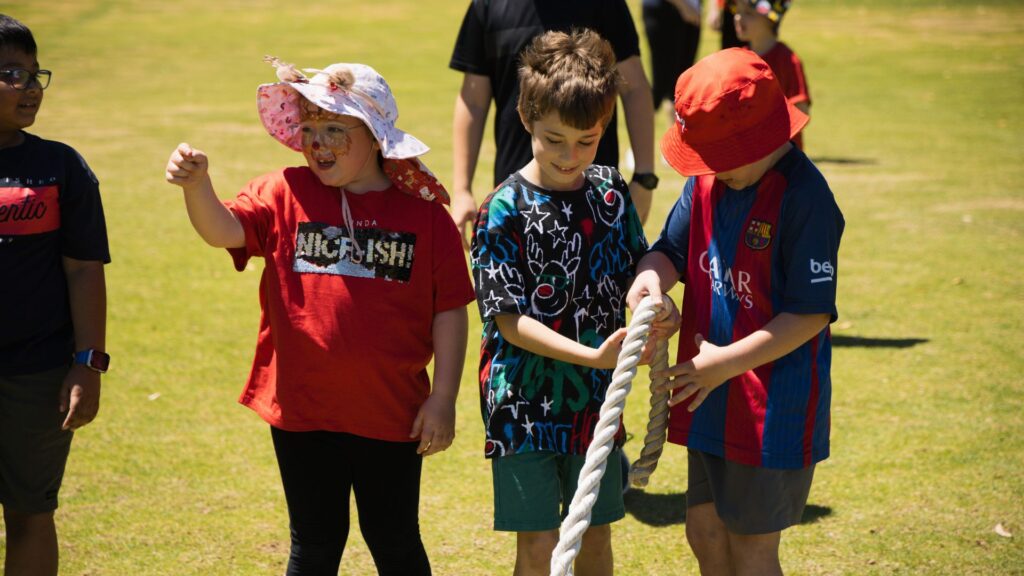 Three children outside, wearing hats and exercise gear. Playing cooperatively with a large rope on the grass.