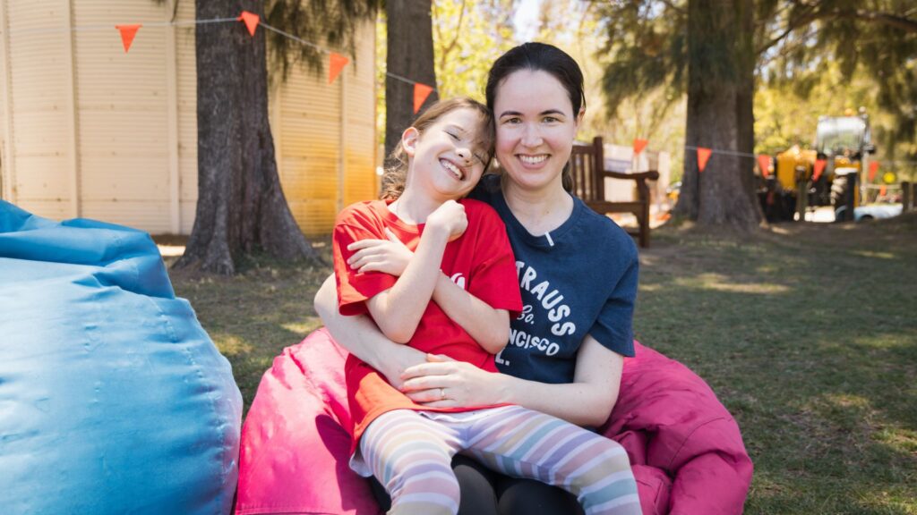 Child sitting on mothers lap. Mother is sitting on a pink beanbag and both are smiling at the camera.