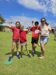 Three children and an adult in Thriving in Motion shirts flexing and celebrating together on a grass oval on a sunny day.