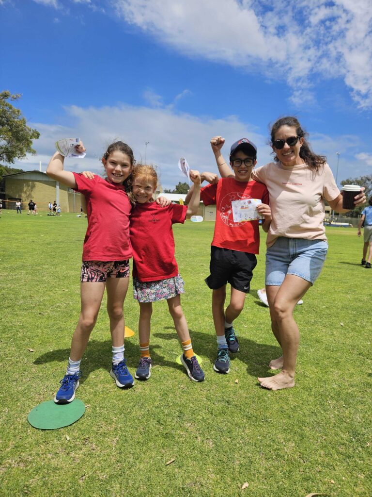 Three children and an adult in Thriving in Motion shirts flexing and celebrating together on a grass oval on a sunny day.