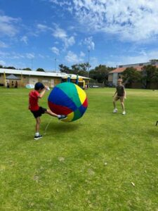 A child in a red Thriving in Motion shirt kicking a giant colourful ball on a grass oval at a community event.