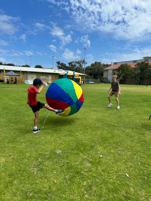 A child in a red Thriving in Motion shirt kicking a giant colourful ball on a grass oval at a community event.