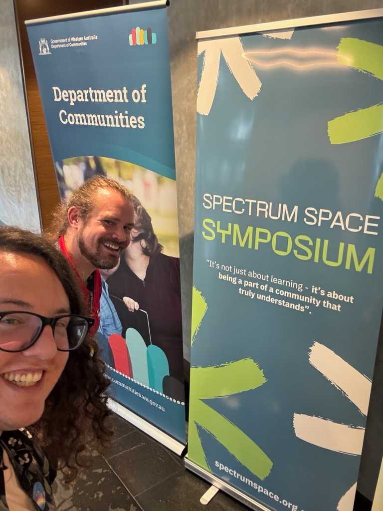 Ben Kramer and Ben Quick from Thriving in Motion at the Spectrum Space Symposium, standing beside the Department of Communities banner
