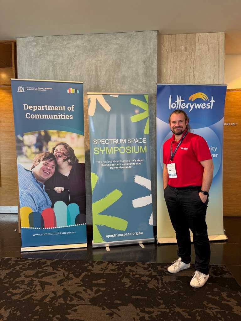 Ben Kramer, Business Manager at Thriving in Motion, standing beside the Spectrum Space Symposium and Lotterywest banners at the conference