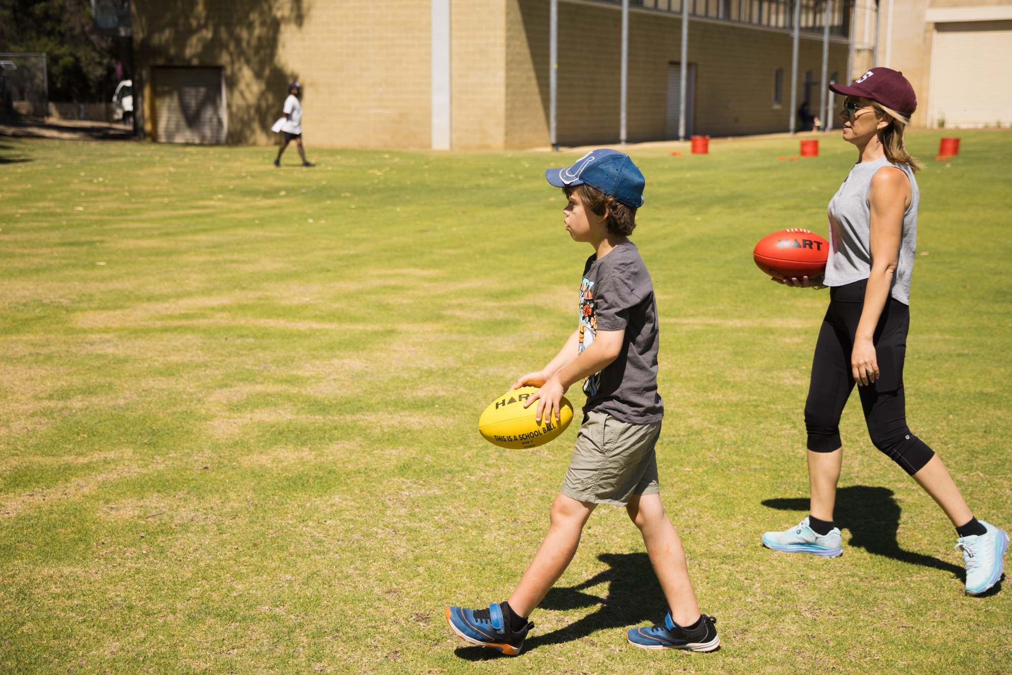 A child and an adult walking together across a grass oval, each carrying a football