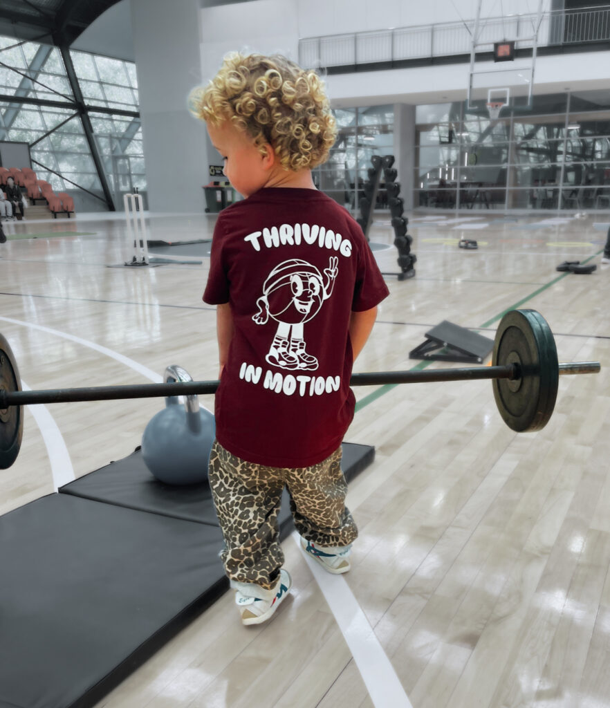 small toddler holding onto a weighted barbell wearing a Thriving in Motion top