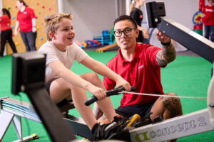 An exercise physiologist coaching a boy on a rowing machine, both smiling.